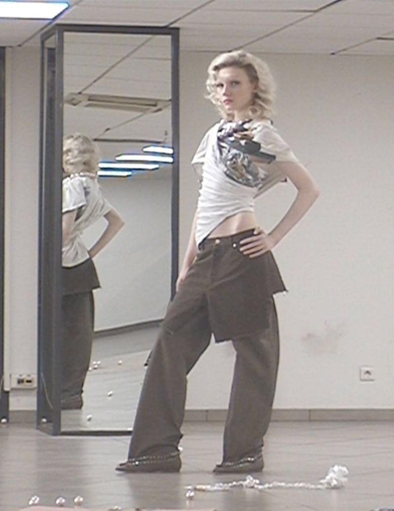 A slender fashion model poses in a casual outfit in an indoor setting, reflected in a mirror behind, accented by scattered pearls.