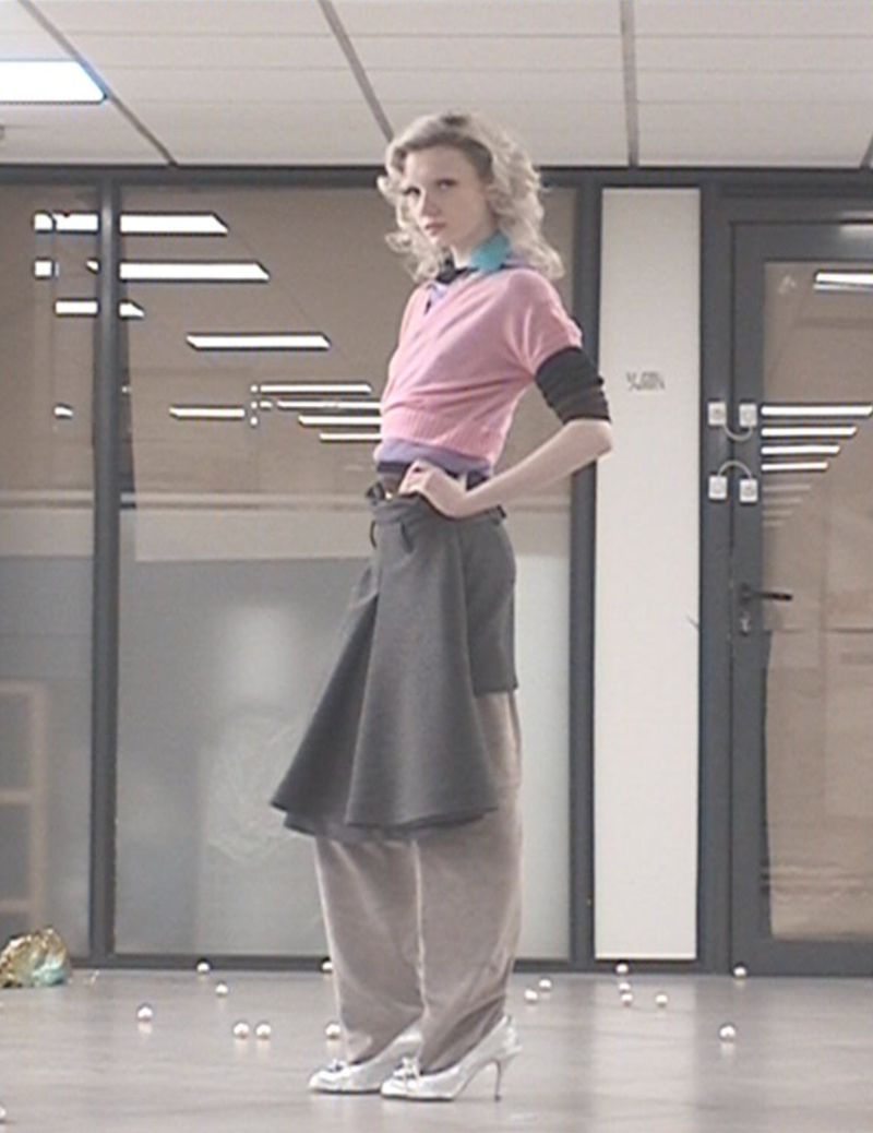 Adult female fashion model in layered top and gray skirt posing indoors against glass doors and scattered balls on the floor.