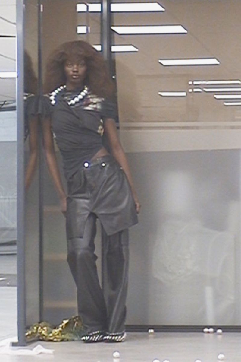 A dark-skinned female model poses in a black top, grey pants, and pearl jewelry, leaning against a partition in a neutral indoor setting.