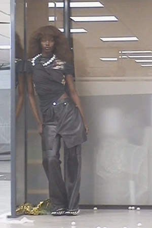 A dark-skinned female model poses in a black top, grey pants, and pearl jewelry, leaning against a partition in a neutral indoor setting.