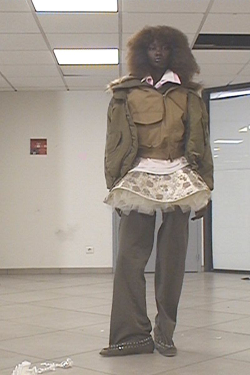 Fashionable woman in a cropped puffer jacket, mini skirt, and loose grey pants poses in an indoor setting with overhead lighting.