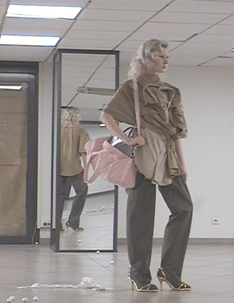 Female model in a studio setting with a large pink bag, wearing an oversized khaki top, baggy gray pants, and gold high heels, posing near a mirror.