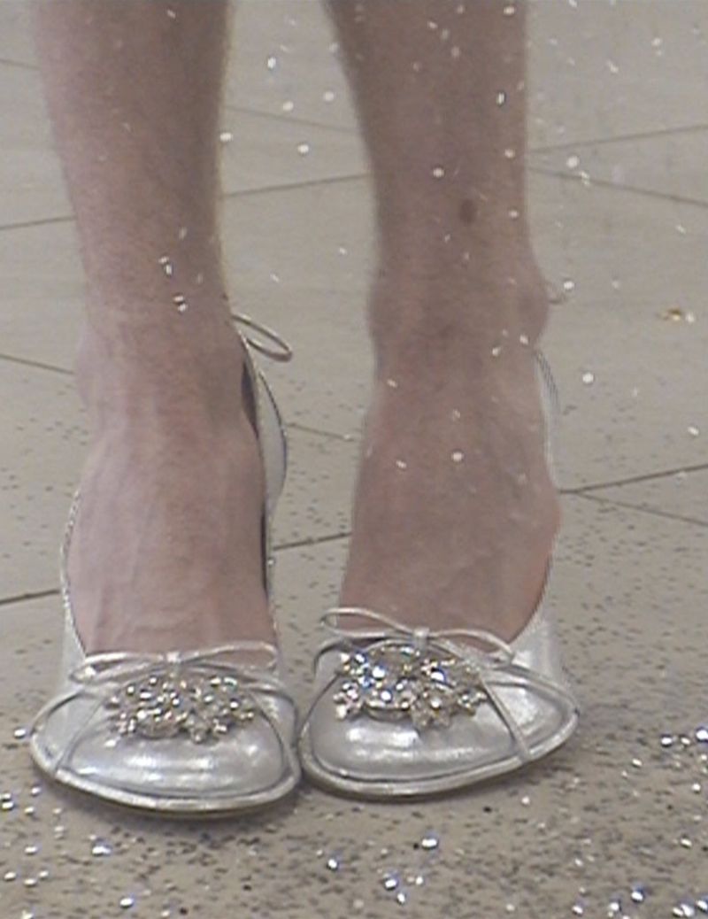 Close-up shot of a person's hairy legs and feet wearing silver ballet shoes with jeweled embellishments, standing on a speckled surface.