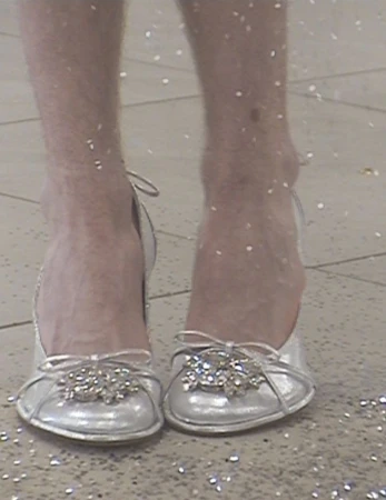 Close-up shot of a person's hairy legs and feet wearing silver ballet shoes with jeweled embellishments, standing on a speckled surface.