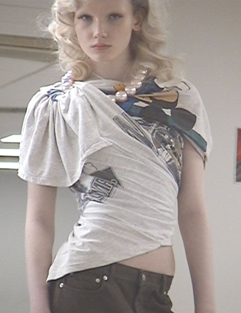 Young adult fashion model poses indoors wearing a repurposed grey graphic t-shirt, brown pants, and a large faux pearl necklace.