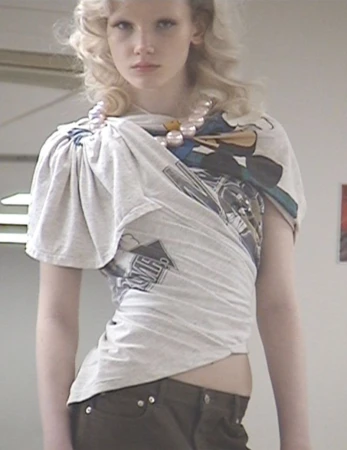 Young adult fashion model poses indoors wearing a repurposed grey graphic t-shirt, brown pants, and a large faux pearl necklace.