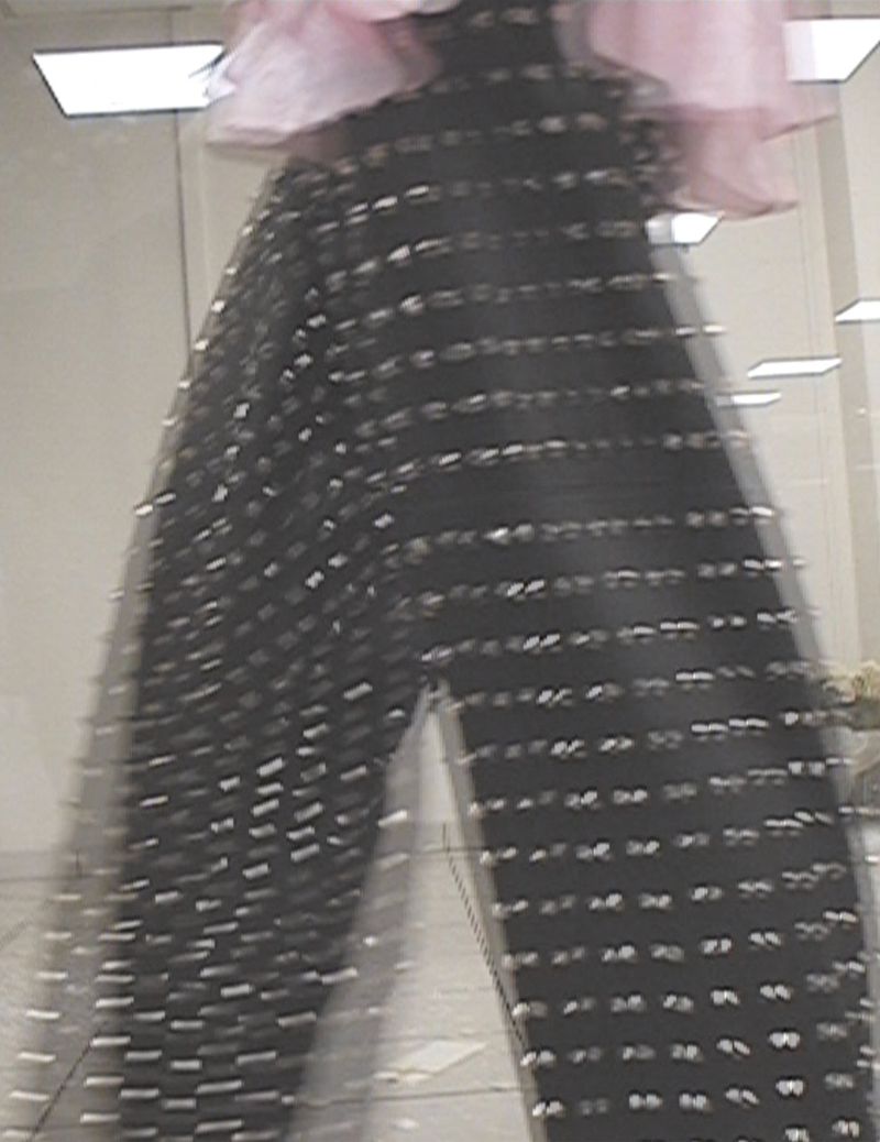 Fashion shot: A person wears black mesh pants adorned with shiny accents, paired with a light pink top, against a neutral interior backdrop.