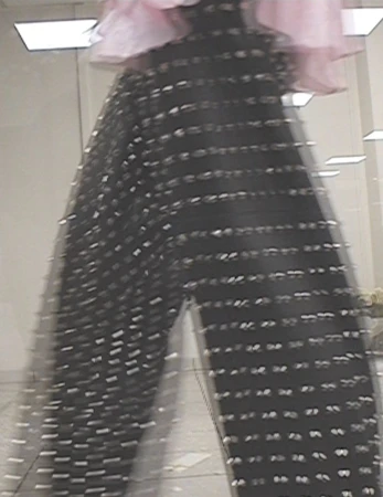 Fashion shot: A person wears black mesh pants adorned with shiny accents, paired with a light pink top, against a neutral interior backdrop.
