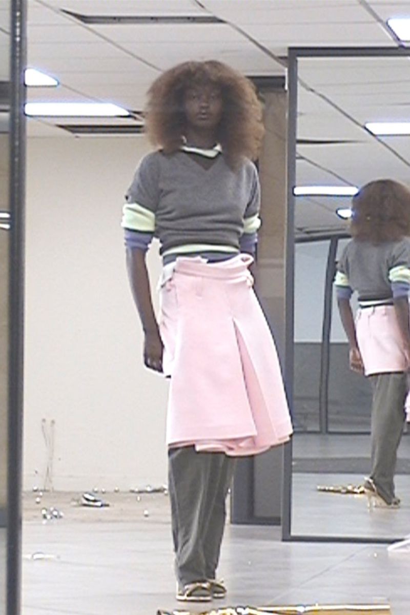 Model with voluminous hair wearing a grey sweater, layered pink skirt, and grey trousers in an indoor space with mirrors.