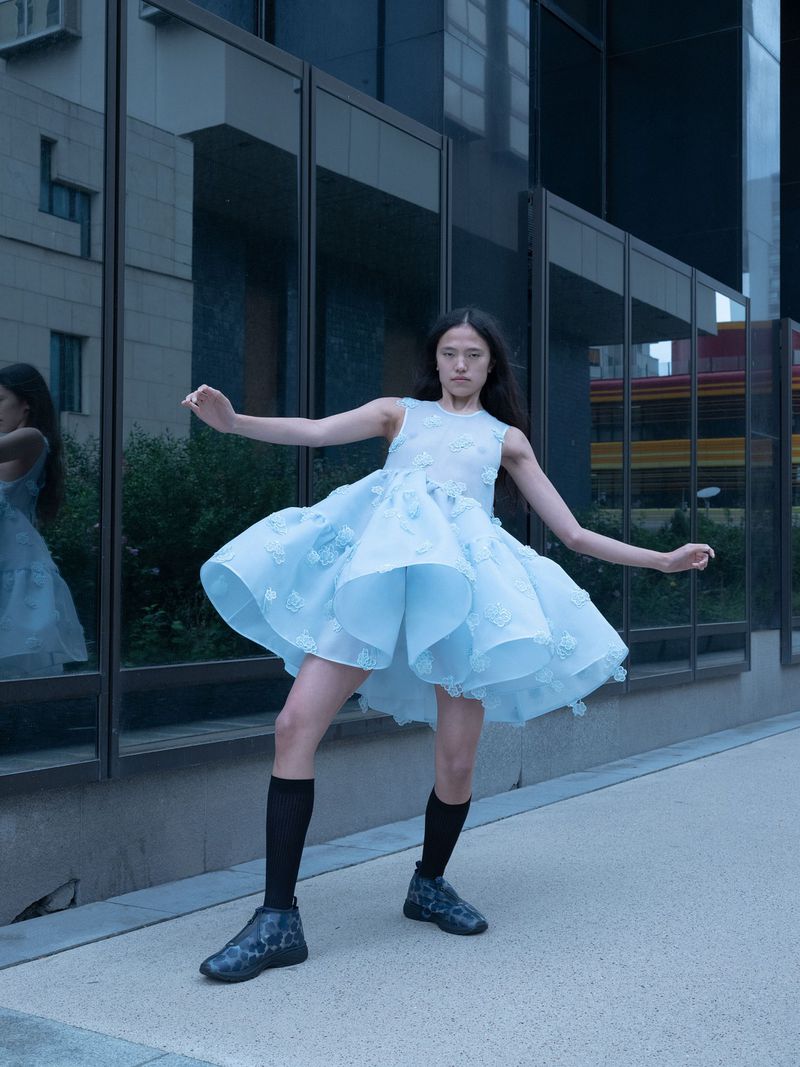 Young woman in a light blue dress with floral accents poses in an urban setting with glass buildings, showcasing fashion and modern style.