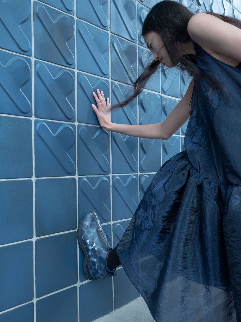A woman with long, dark hair poses with her hand on a blue-tiled wall while wearing a flowing dress and patterned shoe, presenting a fashion-editorial look.