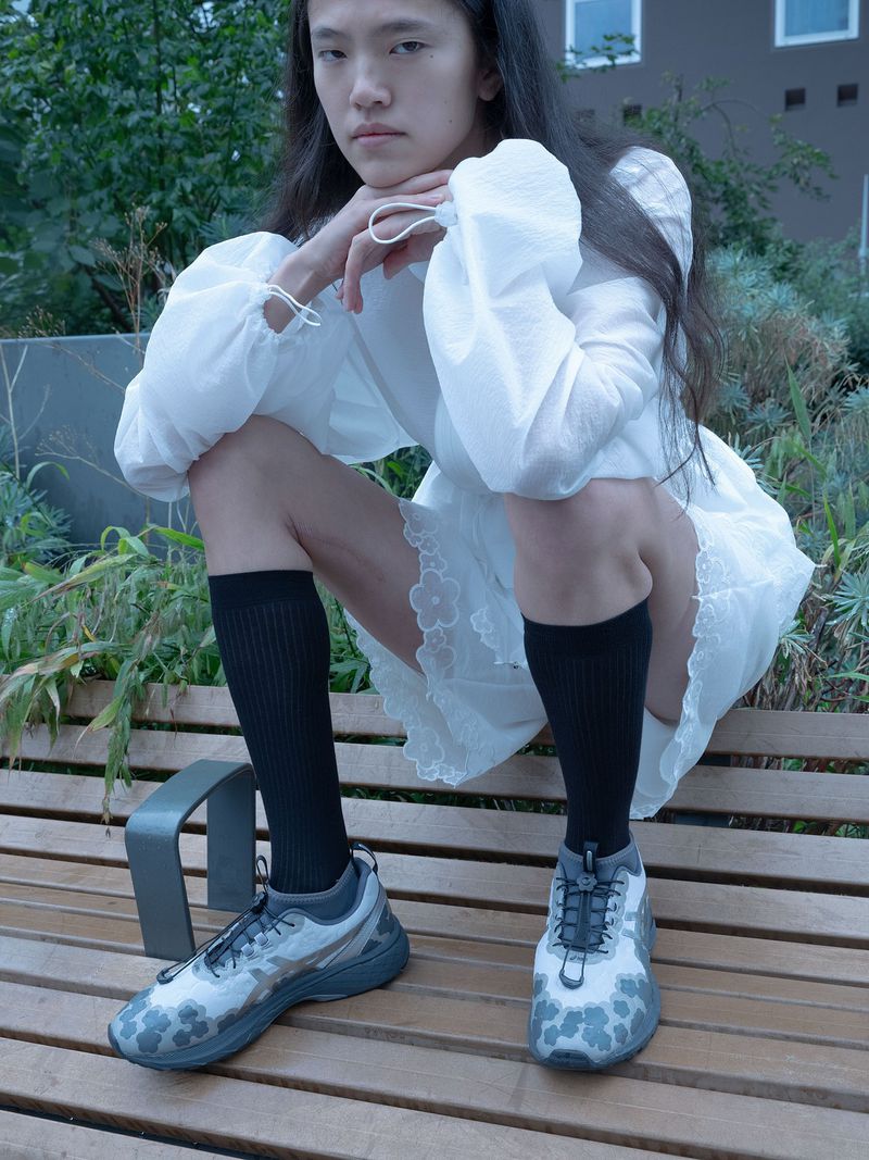 An adult posing outdoors on a wood bench wearing black socks and grey running shoes. The subject wears a white mini-dress and has long dark hair.