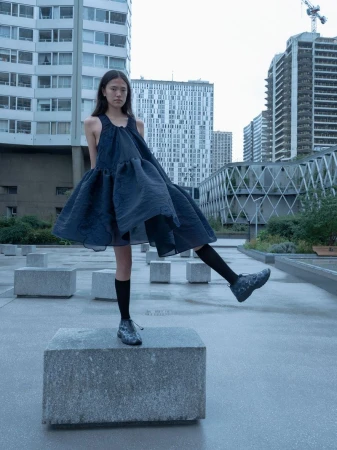Young woman poses in a dark dress and socks, standing on a concrete block in an urban environment with tall buildings visible.