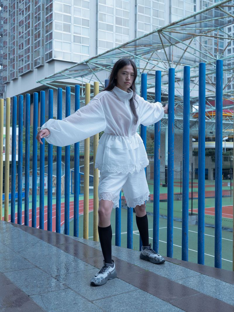 A young adult woman models translucent white fashion outdoors, standing on a gray ledge against a blue and yellow fence and cityscape backdrop.