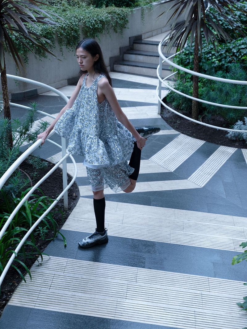 Young Asian woman in shimmering blue dress stretches leg on walkway with greenery. Stairs and metal railings add architectural detail.