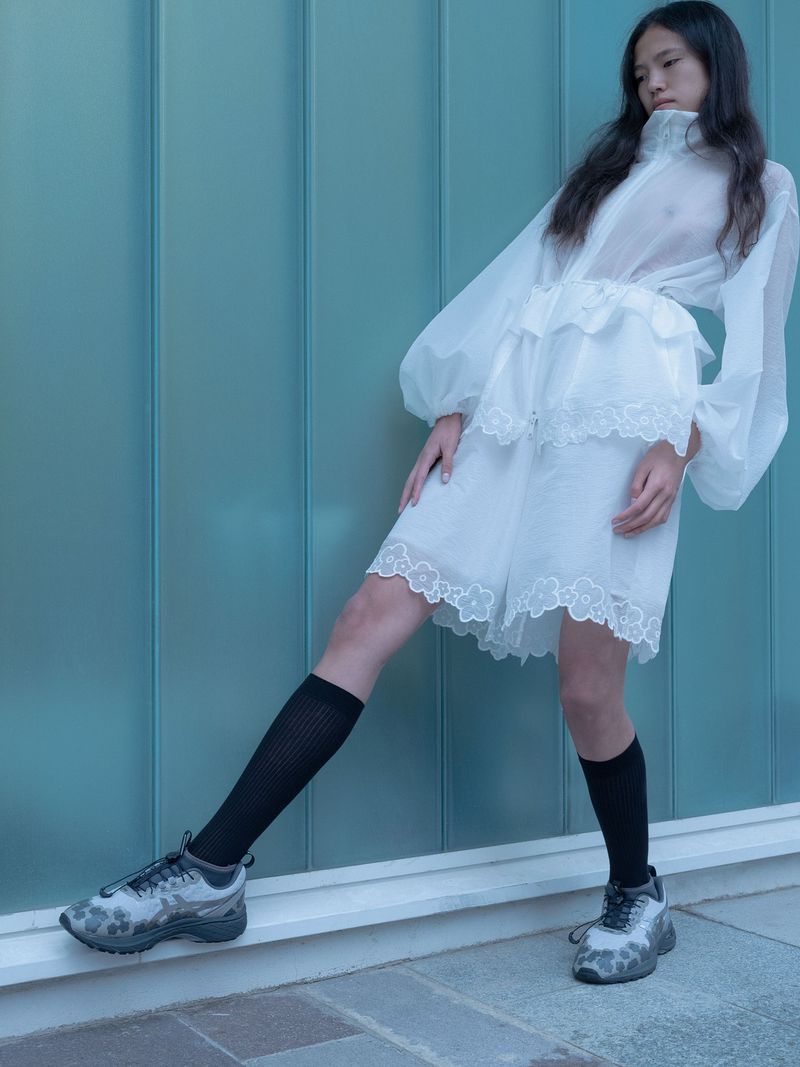 Model in white sheer dress and black socks wearing patterned sneakers against a teal wall