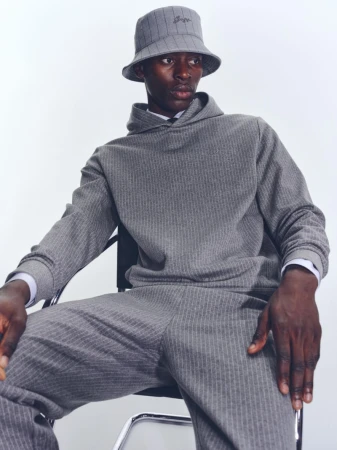 Model wearing a grey striped tracksuit and matching bucket hat seated on a chair against a white background.