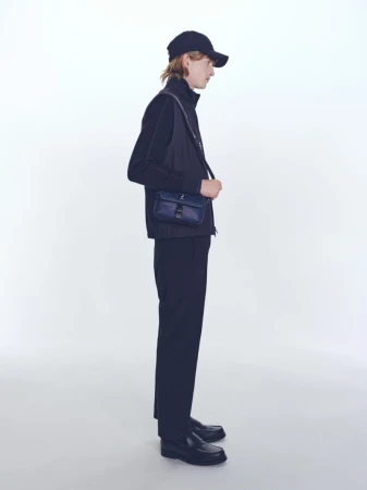 Profile shot of a young adult male model in black casual wear with a navy messenger bag against a plain white backdrop.