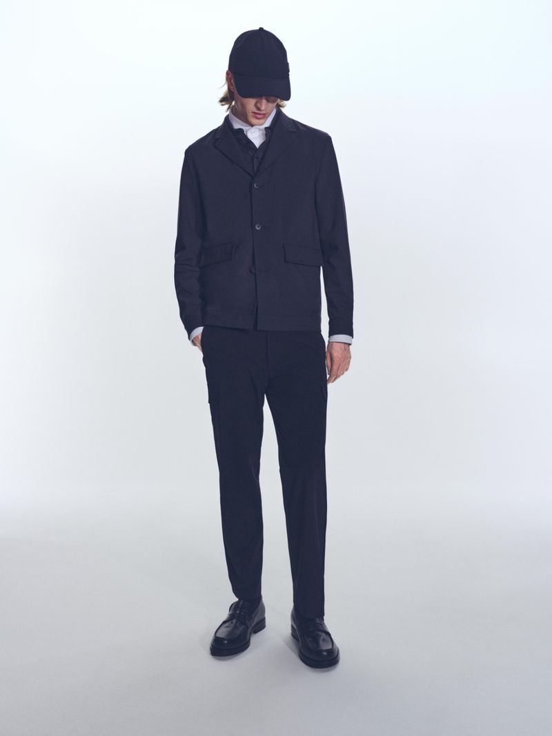 A slender man in a blazer, dress shirt, dark trousers, and cap stands on a white background in a studio shot, fashion editorial.