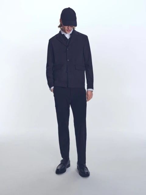 A slender man in a blazer, dress shirt, dark trousers, and cap stands on a white background in a studio shot, fashion editorial.