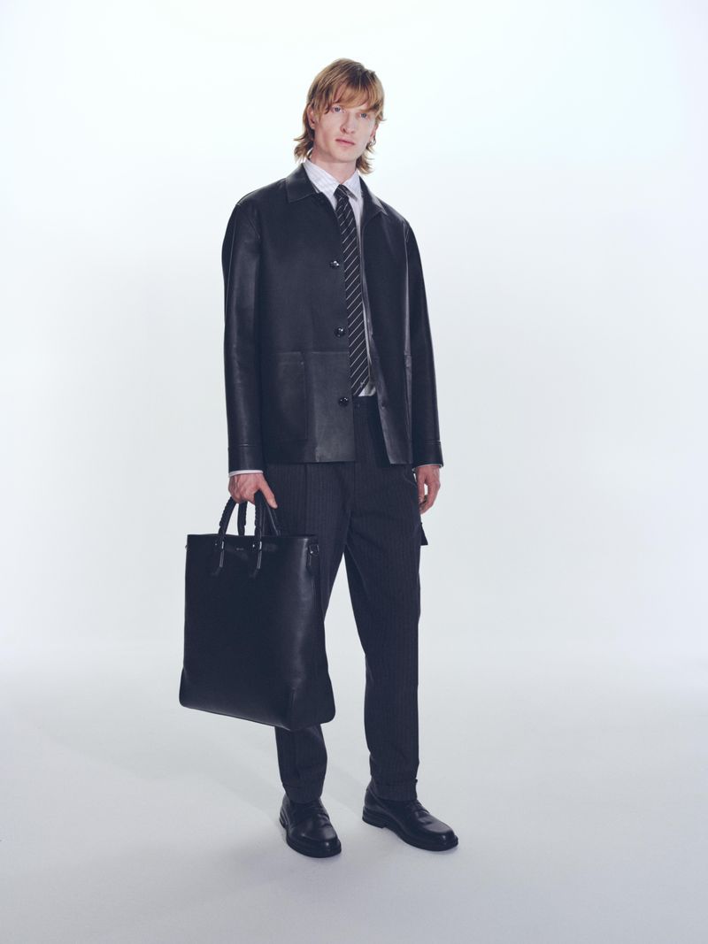 Full-length studio shot of an adult male model wearing a leather jacket, black suit, tie, and shoes, holding a black leather tote bag.