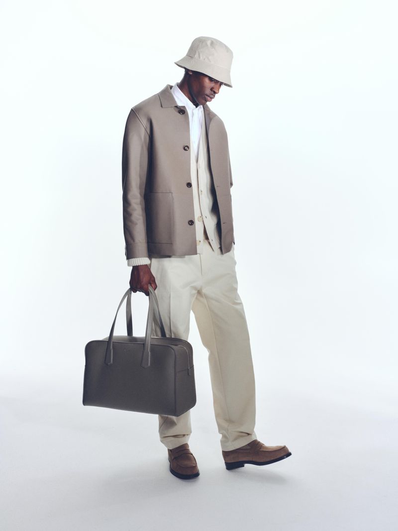Dark-skinned adult male in light-brown suit, bucket hat, and brown loafers standing in a bright, neutral studio and holding a bag.