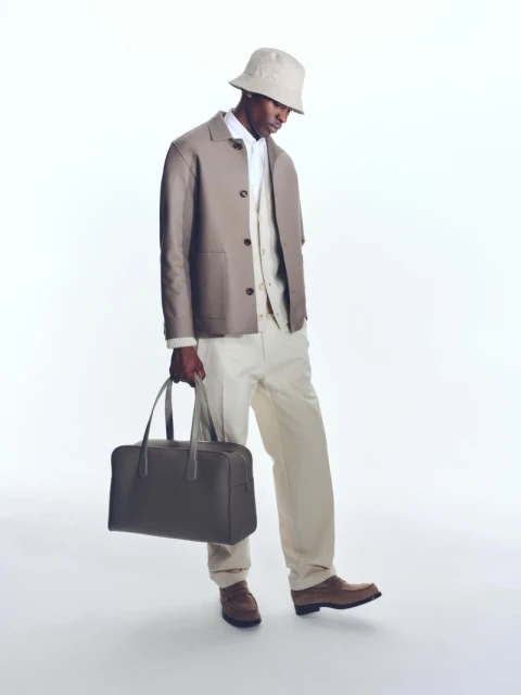 Dark-skinned adult male in light-brown suit, bucket hat, and brown loafers standing in a bright, neutral studio and holding a bag.