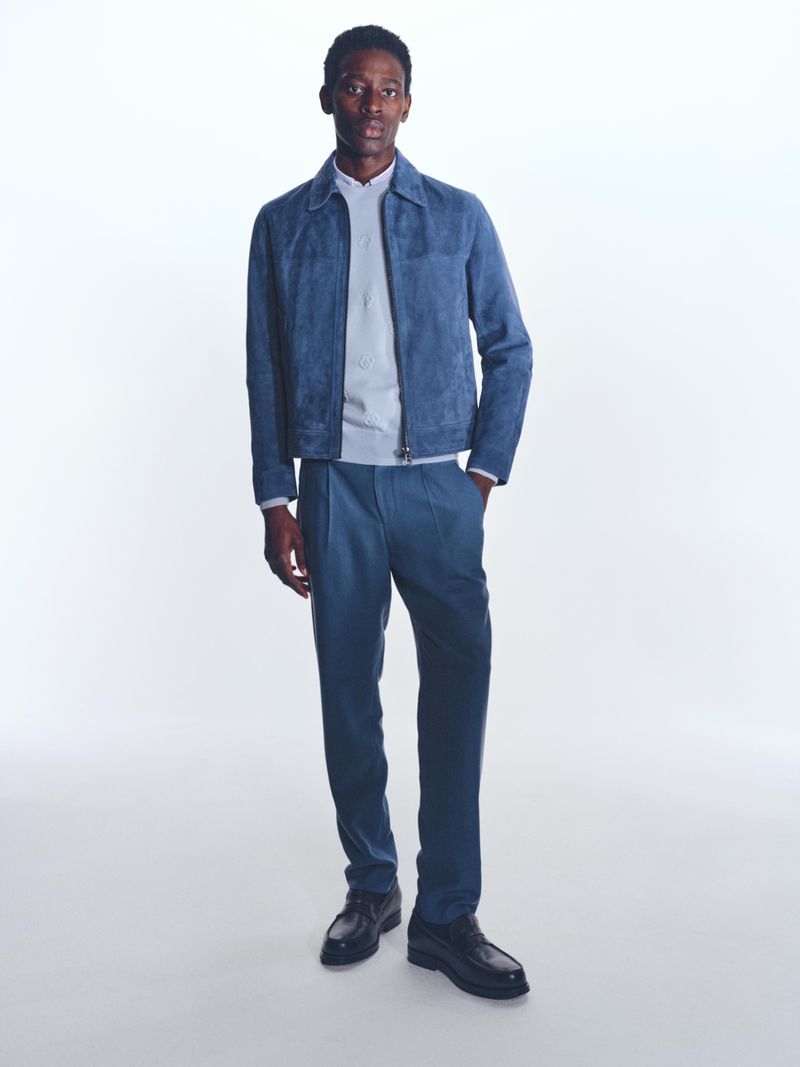 Full-length studio shot of a Black male model wearing a blue suede jacket, sweater, pants, and dress shoes against a white backdrop.