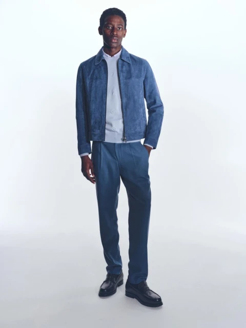 Full-length studio shot of a Black male model wearing a blue suede jacket, sweater, pants, and dress shoes against a white backdrop.