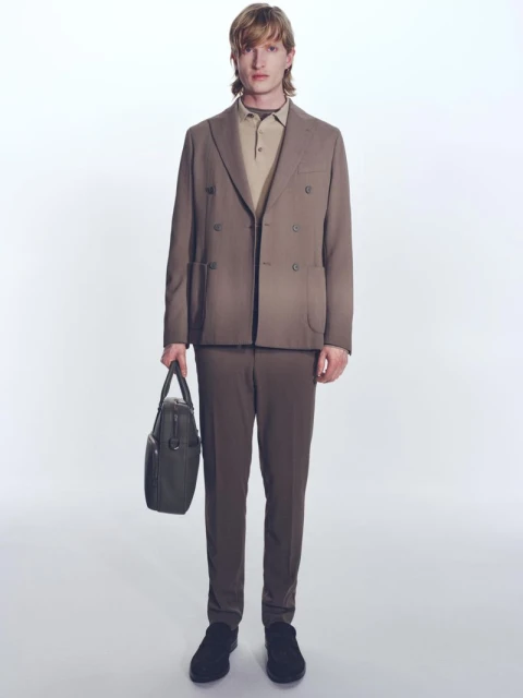 Adult male in a brown suit and polo shirt holds a black briefcase, standing in a brightly lit photo studio. Fashion editorial shot.