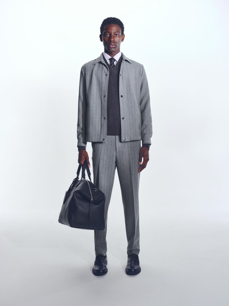 Black man in a grey pinstripe suit holding a black leather duffel bag, posing in a bright photo studio, looking at camera.