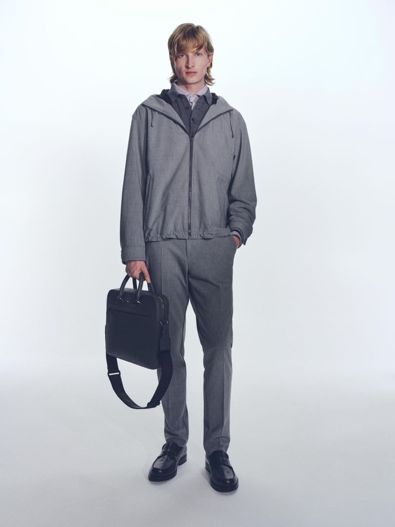 A slender adult male model poses in a grey hooded jacket, trousers, shirt, and loafers. He is holding a black leather business bag.