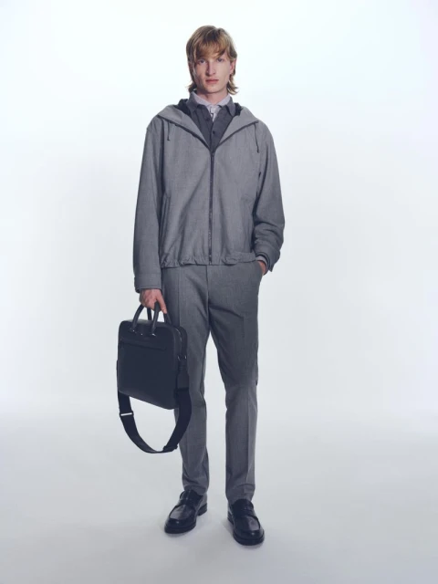 A slender adult male model poses in a grey hooded jacket, trousers, shirt, and loafers. He is holding a black leather business bag.