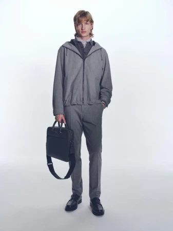 A slender adult male model poses in a grey hooded jacket, trousers, shirt, and loafers. He is holding a black leather business bag.