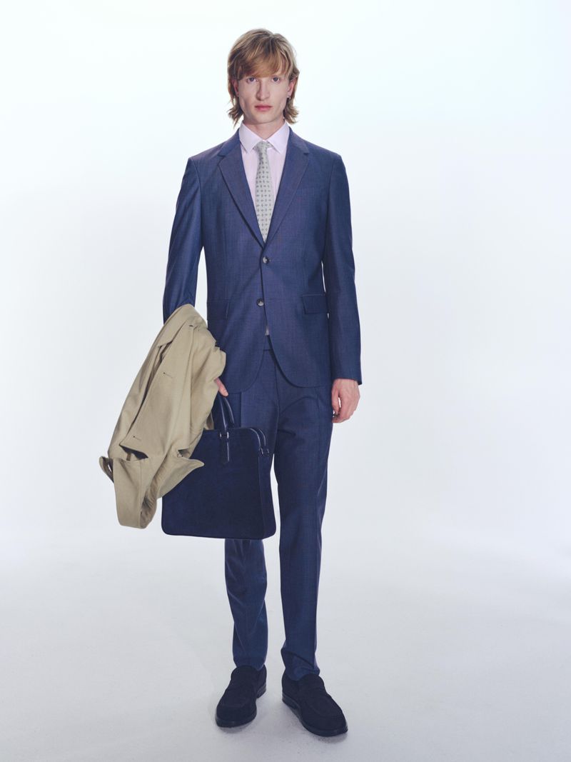 Man in blue suit standing in a bright white studio, holding a beige overcoat and black briefcase in hand, professional attire.