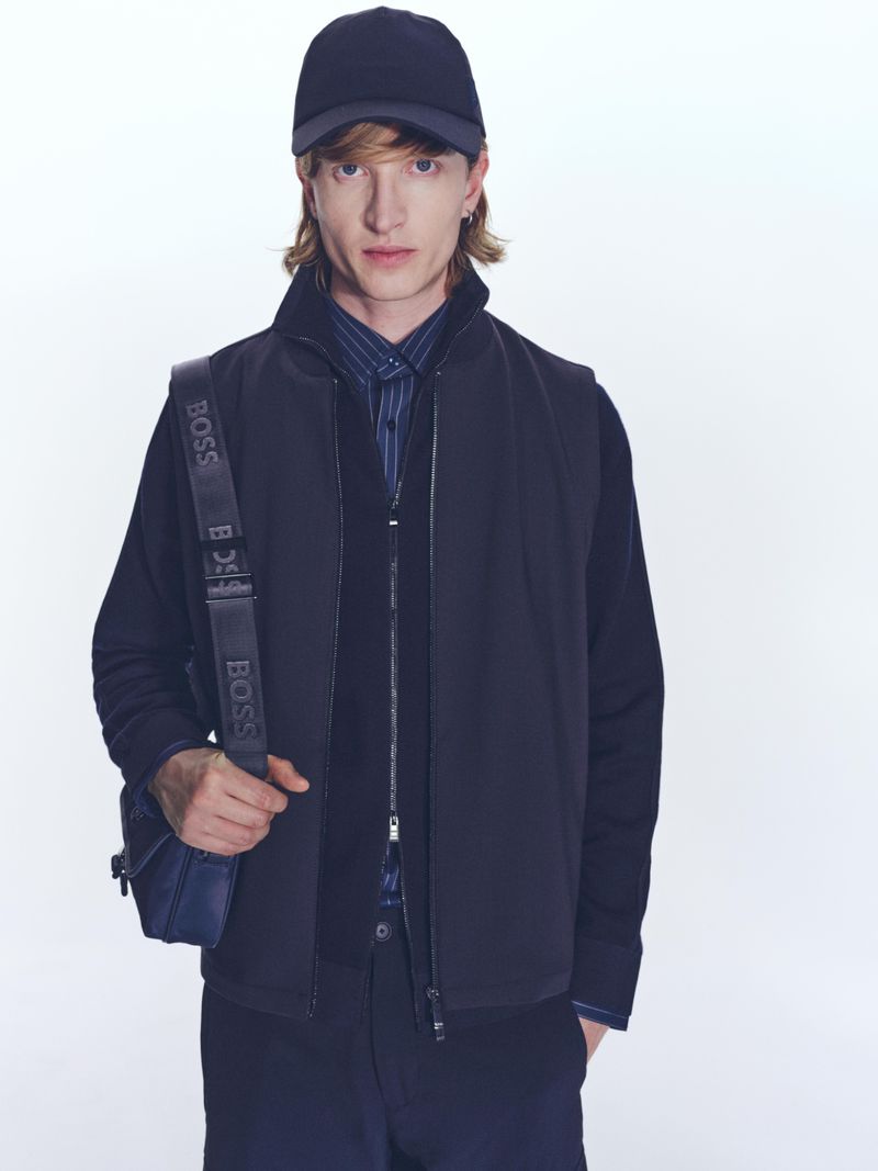 A fashionable adult man wearing a navy cap and vest over a button-down shirt, stands in a photo studio with a neutral backdrop.