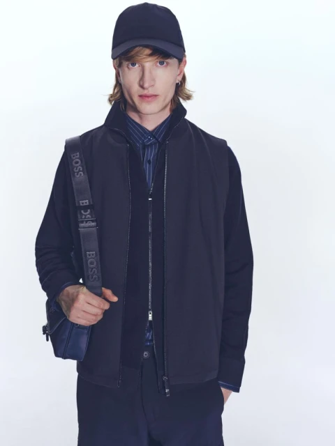 A fashionable adult man wearing a navy cap and vest over a button-down shirt, stands in a photo studio with a neutral backdrop.