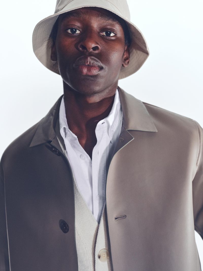 Portrait of a Black male model in a beige bucket hat, white shirt, and beige coat, set against a bright white studio backdrop.