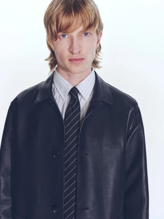 Model wearing a black leather jacket, striped shirt, and dark tie against a clean white background