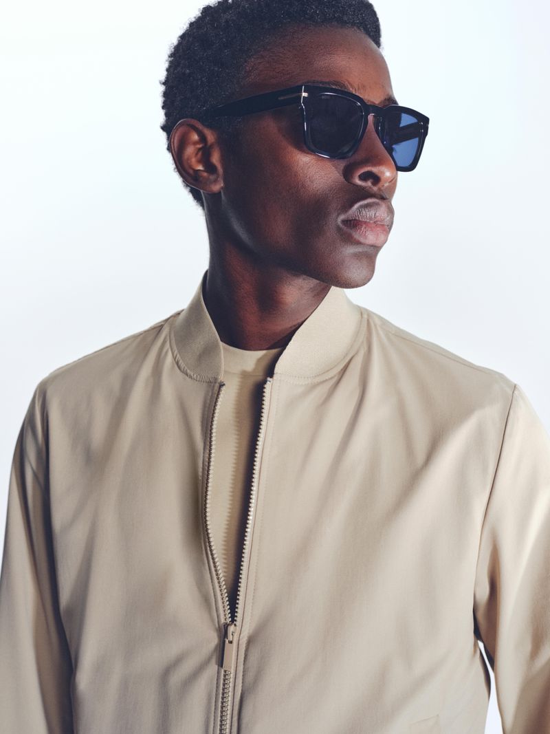 Close-up of a Black male model wearing sunglasses and a beige bomber jacket against a white background in a studio setting.
