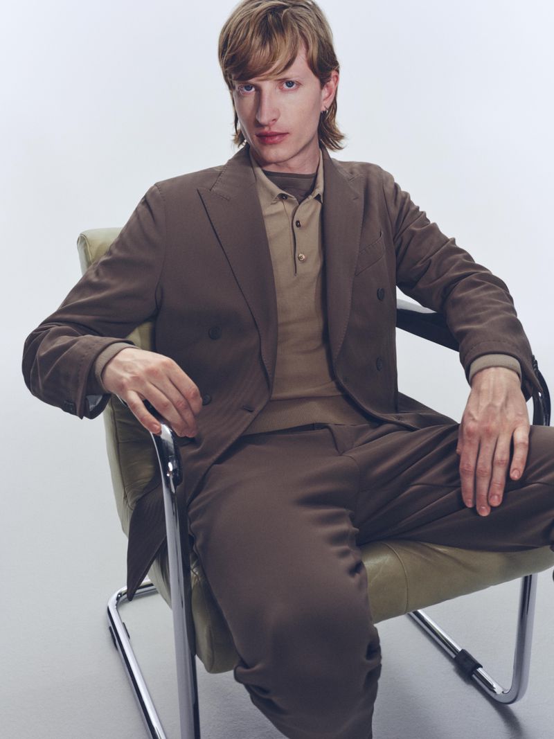 An adult male sits in a retro chair wearing a brown double-breasted suit against a white backdrop, captured in a studio shot.