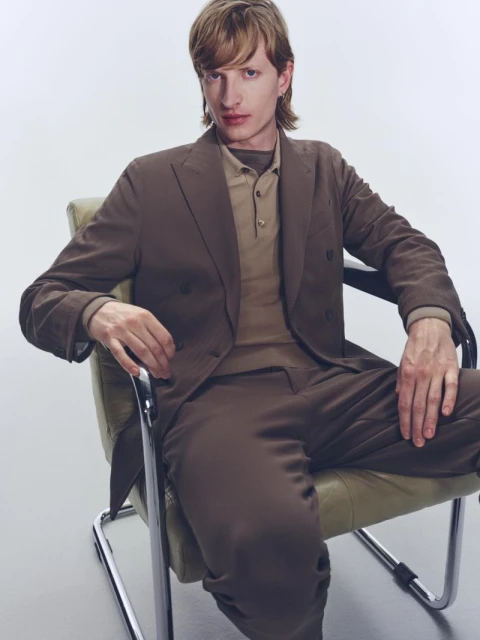 An adult male sits in a retro chair wearing a brown double-breasted suit against a white backdrop, captured in a studio shot.