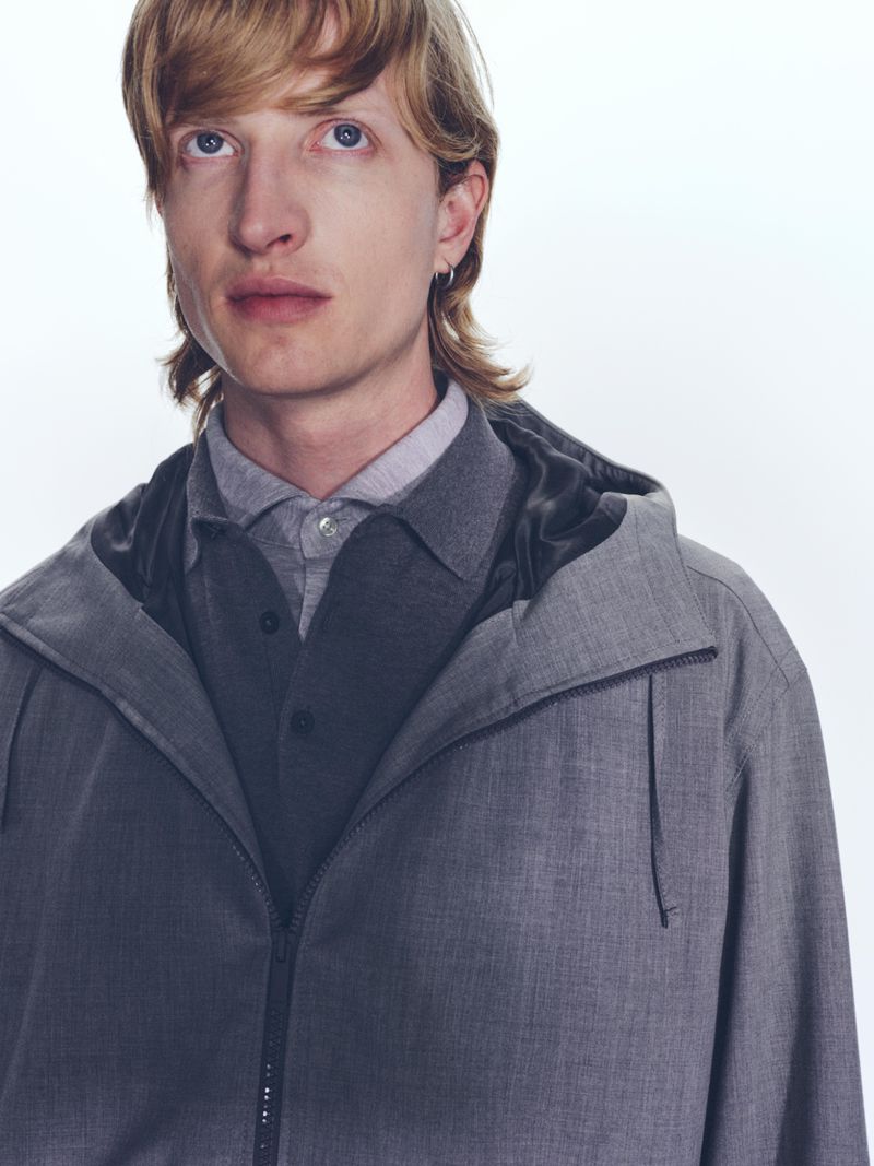Close-up of a blond male model wearing a grey hooded jacket and a grey shirt against a white backdrop in a studio setting.