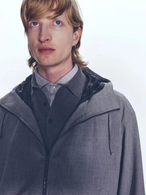 Close-up of a blond male model wearing a grey hooded jacket and a grey shirt against a white backdrop in a studio setting.