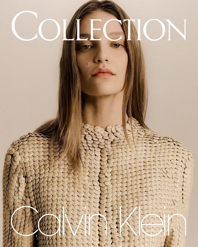 Close-up fashion shot of a woman with light brown hair wearing a beige jacket with a scale-like texture in a neutral studio setting.