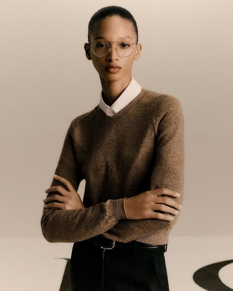 Close-up studio portrait of a fashion model wearing round glasses, a brown sweater over a collared shirt, and black pants.