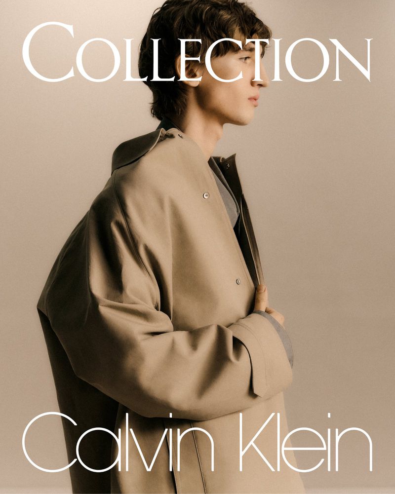 Fashion shot of a young adult man wearing a tan jacket in a studio setting for a Calvin Klein Collection advertisement.