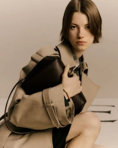 Model in a beige trench coat holding a dark leather shoulder bag against a neutral studio backdrop.