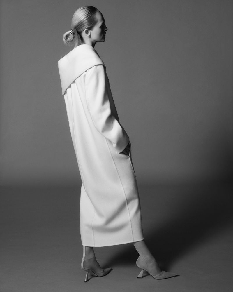 Monochrome profile of a woman in a long white coat and heels against a neutral backdrop showcasing fashion and minimalist style.