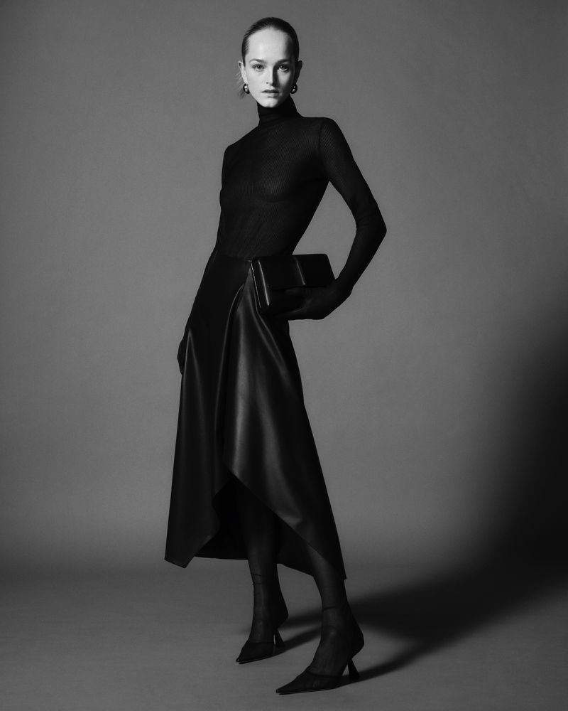 Full-length black-and-white fashion shot: elegant woman with a clutch purse, wearing a sheer top, leather skirt, and high heels.